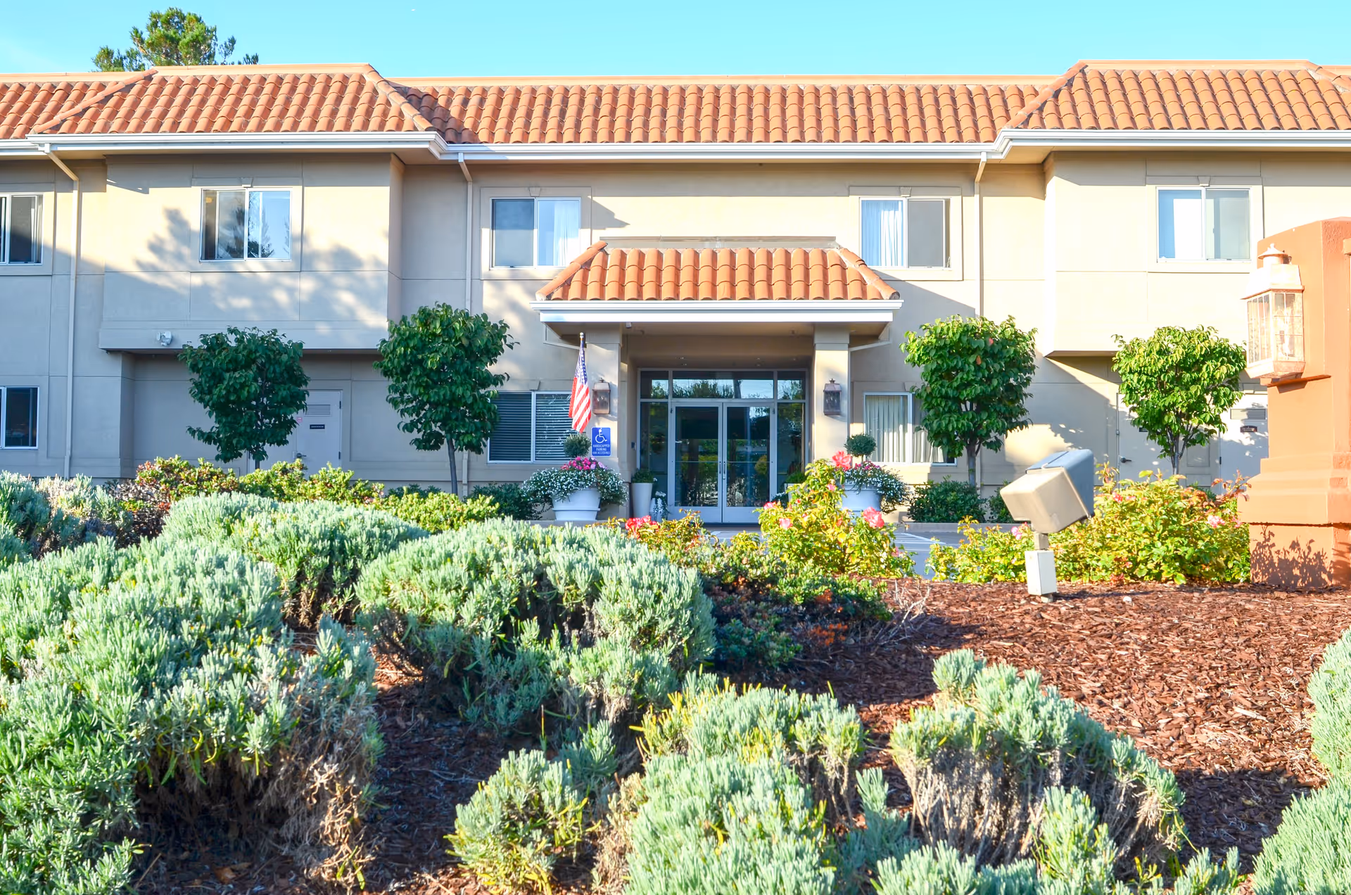 Front exterior view of Mills Estate Villa, a two-story building with a tiled roof, large glass entrance doors, and landscaped greenery including bushes and small trees in the foreground.