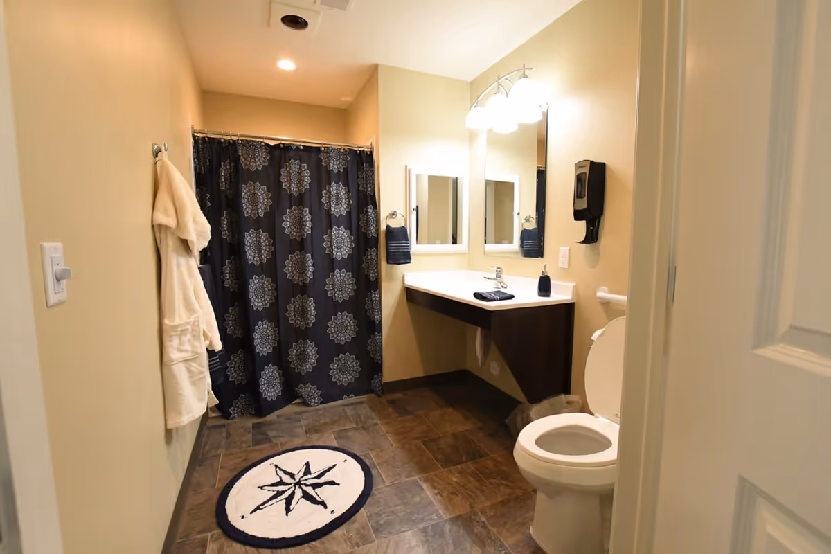 A clean and well-lit bathroom with a toilet, a wall-mounted sink with a large mirror above it, a soap dispenser, a towel ring with a dark towel, a shower area with a dark patterned curtain, a white bathrobe hanging on the wall, and a round rug with a compass design on the floor.