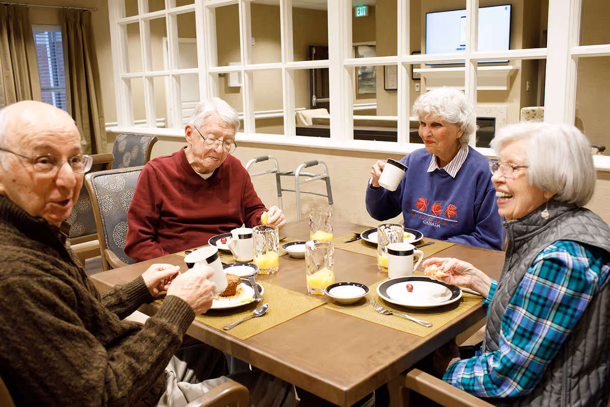 Four elderly residents seated around a dining table enjoying coffee and breakfast in a communal dining room.
