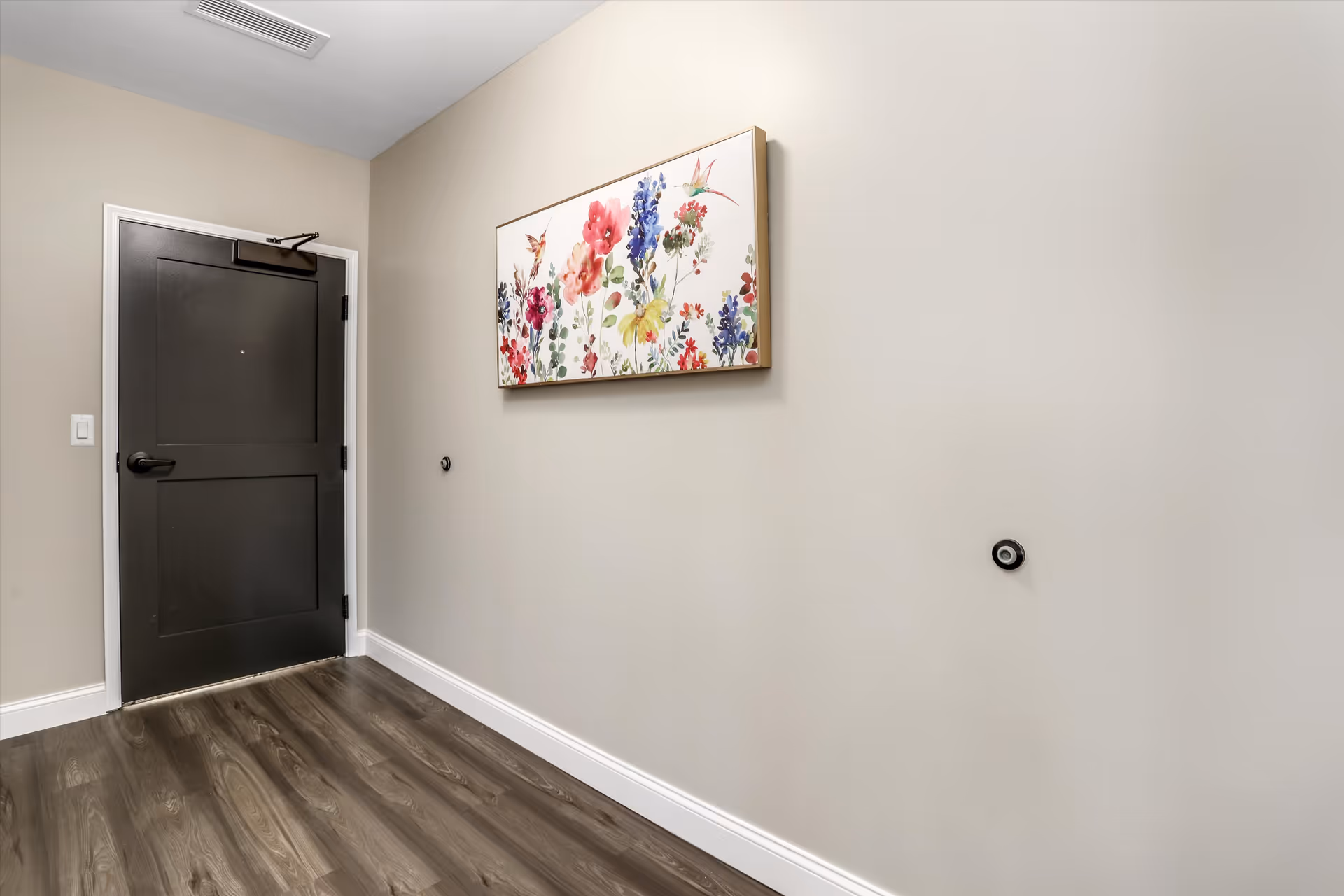 An entryway with a dark door, wood-look flooring, neutral walls and a colorful floral painting on the wall.
