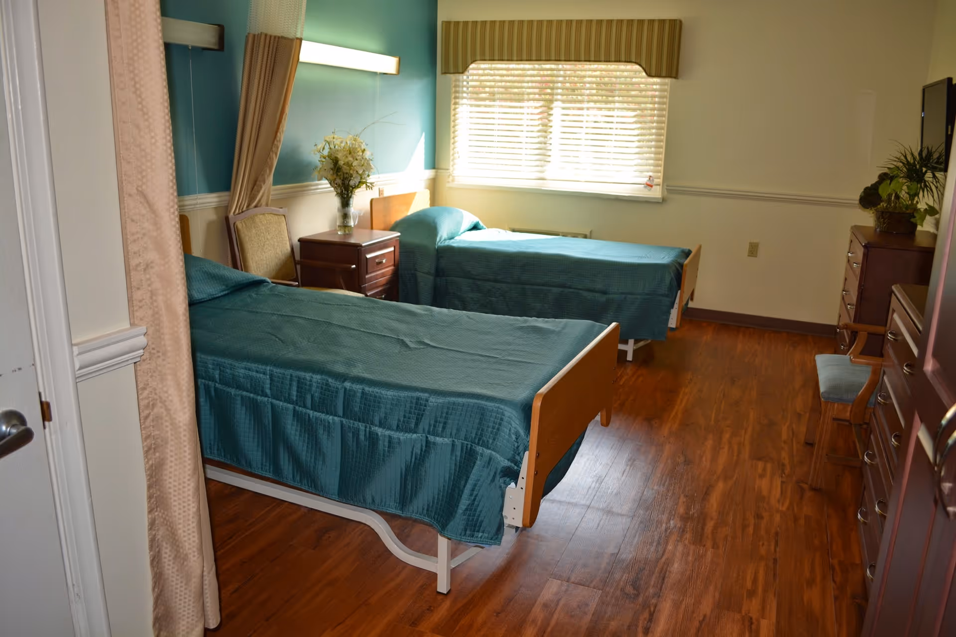 A senior living facility bedroom with two single beds covered in teal bedspreads. The room has wooden flooring, a window with blinds and a striped valance, a nightstand with a vase of flowers, a chair, a dresser with a plant, and a TV mounted on the wall.