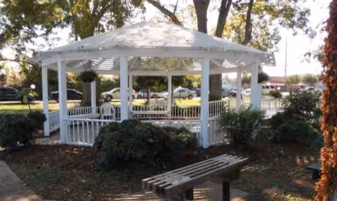 White gazebo in a landscaped outdoor area with a bench and shrubs.
