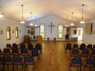 Interior view of a chapel or worship room with rows of chairs facing a small altar area. The room has wooden flooring, several stained glass windows, a large cross mounted on the wall, and pendant lights hanging from the ceiling.
