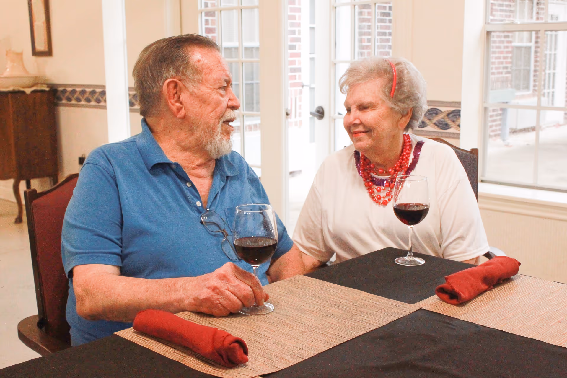 An older man and woman sit at a dining table, smiling at each other while holding glasses of red wine.