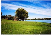 A scenic outdoor view featuring a large green grassy area with a tree and a wooden gazebo near the edge of a calm lake under a partly cloudy blue sky.