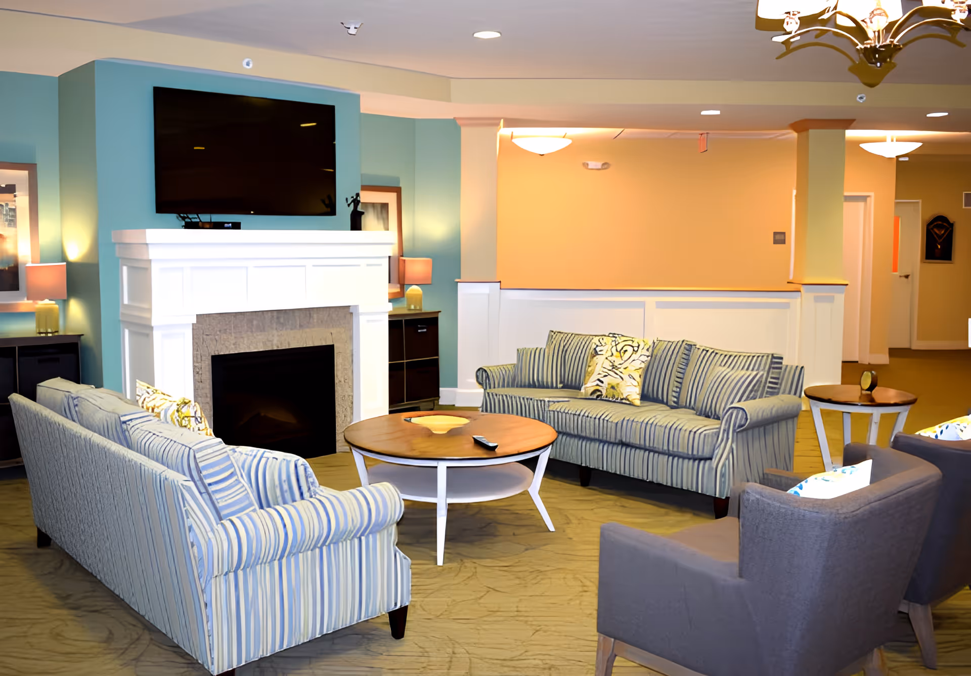 A cozy living room area in Sanctuary Grande featuring two striped sofas, two gray armchairs, a round wooden coffee table, and a fireplace with a mounted flat-screen TV above it. The walls are painted in soft teal and beige tones, with lamps and framed artwork adding to the warm ambiance.