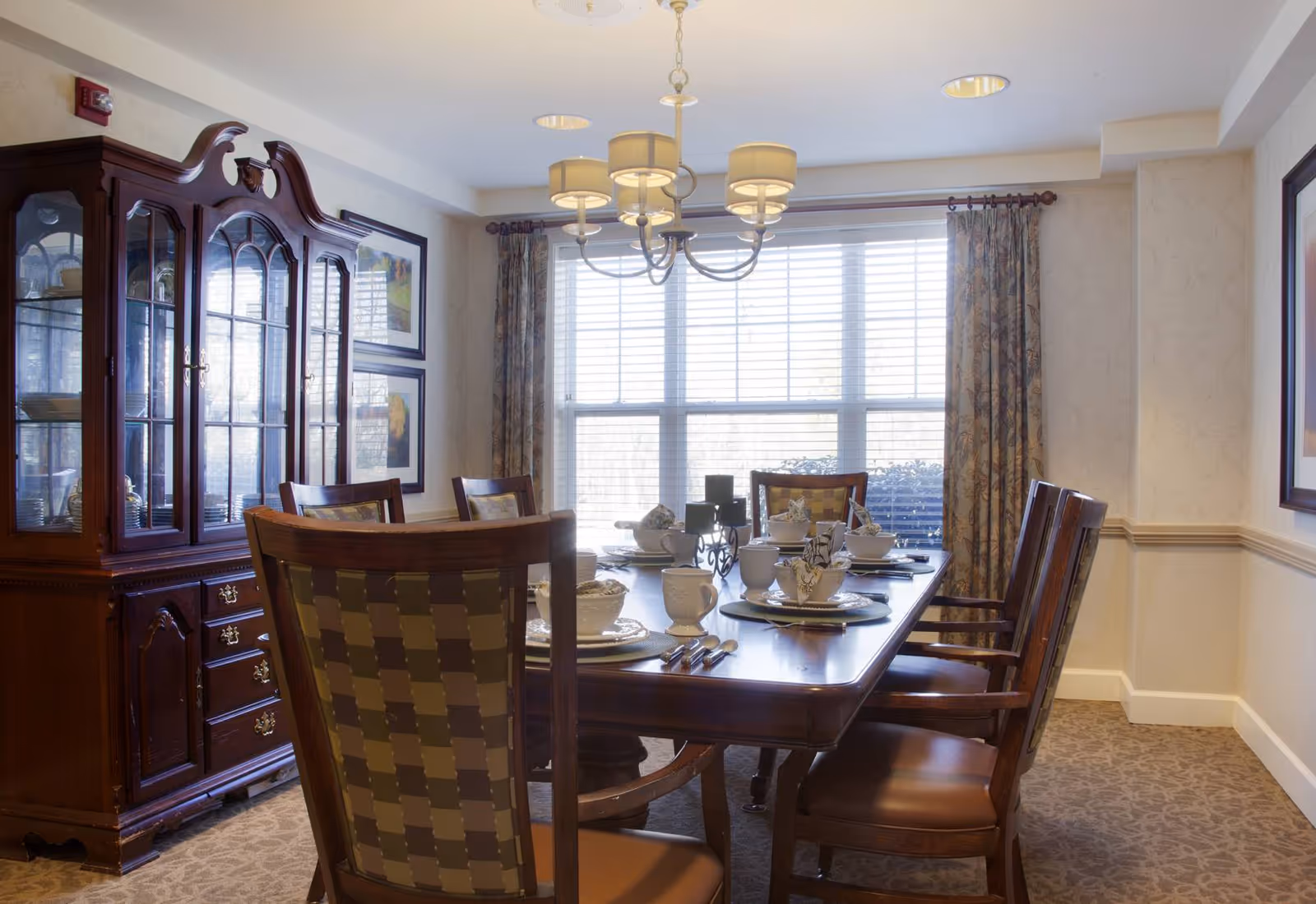 A well-lit dining room with a large wooden dining table set for six people. The table is arranged with plates, cups, and napkins. There is a wooden china cabinet with glass doors on the left side of the room. A chandelier with multiple lampshades hangs above the table. The room has patterned carpet, floral curtains, and framed artwork on the walls.
