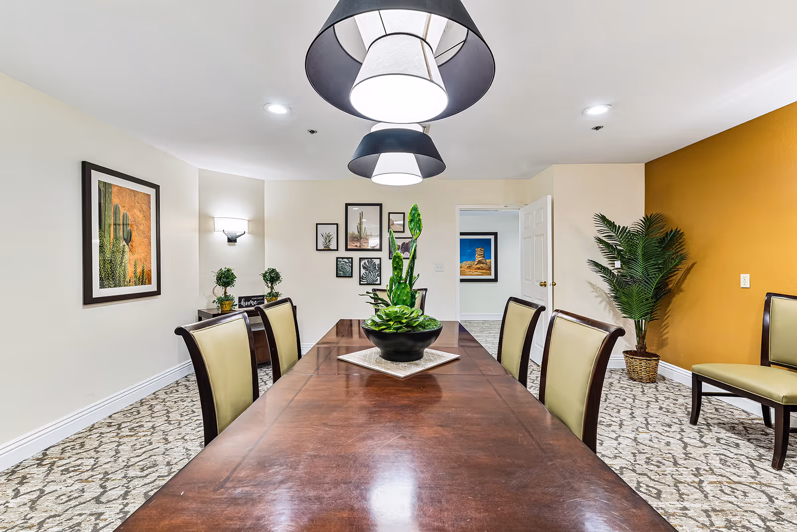 Long wooden dining table with chairs, pendant lights overhead, and decorative plants and artwork in a well-lit room.