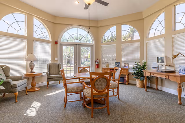 A bright room with large arched windows letting in natural light. The room features a round wooden table with four cushioned chairs in the center, two upholstered armchairs with a small round side table and lamp on the left, and a wooden desk with a vintage computer and a desk lamp on the right. There is a potted plant near the desk and framed photos on the desk and a small cabinet. The room has a carpeted floor and a ceiling fan.