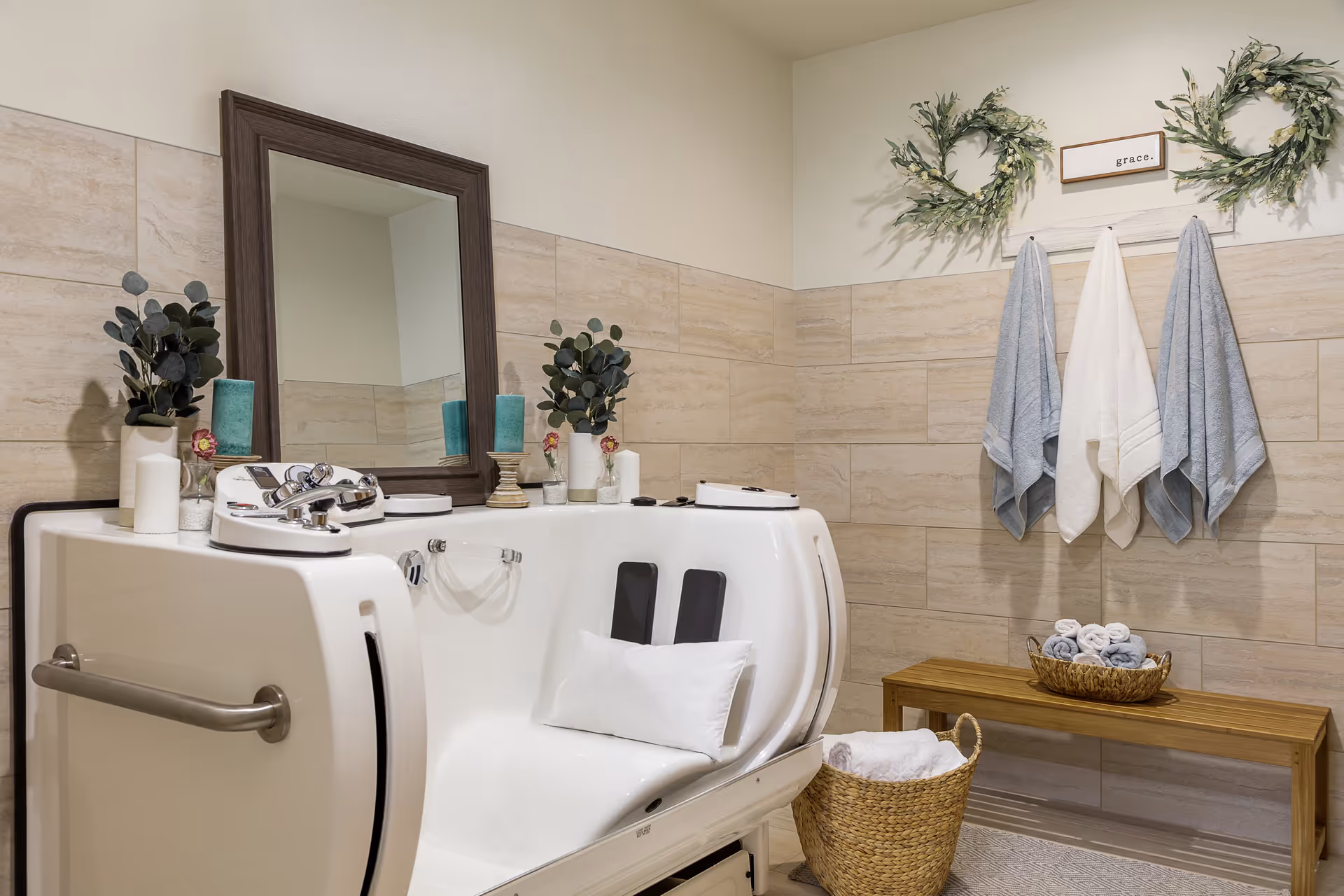 A bathroom with a white walk-in bathtub featuring a pillow and handrails. The walls are tiled with beige tiles, and there is a large mirror above the bathtub. On the wall, there are three towels hanging on hooks beneath two decorative green wreaths and a small sign that says 'grace.' A wooden bench holds a basket with rolled towels, and a woven basket with towels is on the floor.