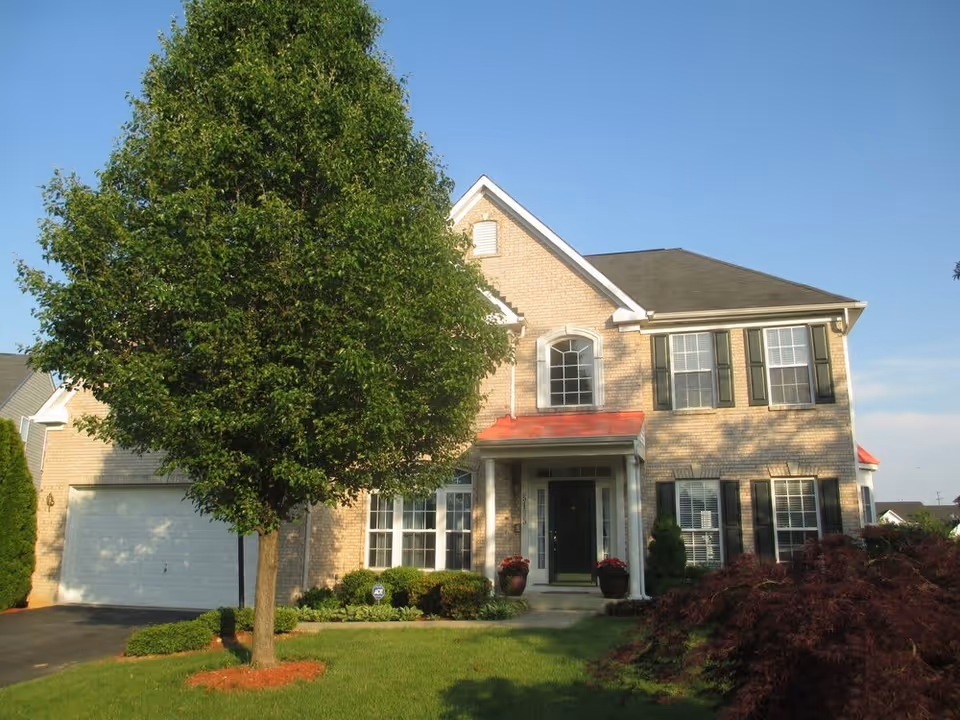A two-story brick house with a front porch, green lawn, and a large tree in the front yard under a clear blue sky.