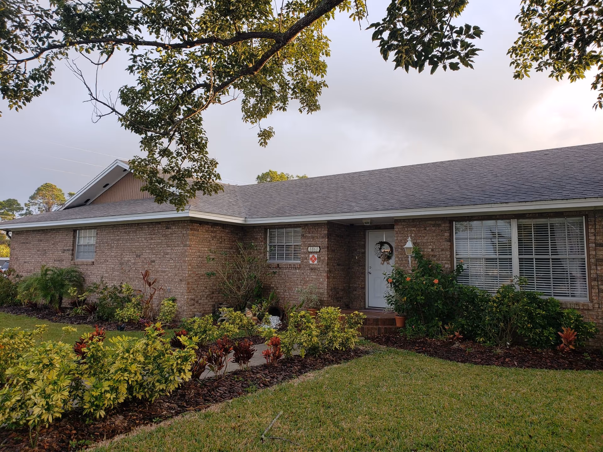 Exterior view of a single-story brick building with a gray shingled roof, surrounded by well-maintained landscaping including bushes and small trees. A white door with a wreath is visible, along with windows with white blinds. The sky is partly cloudy and tree branches frame the top of the image.