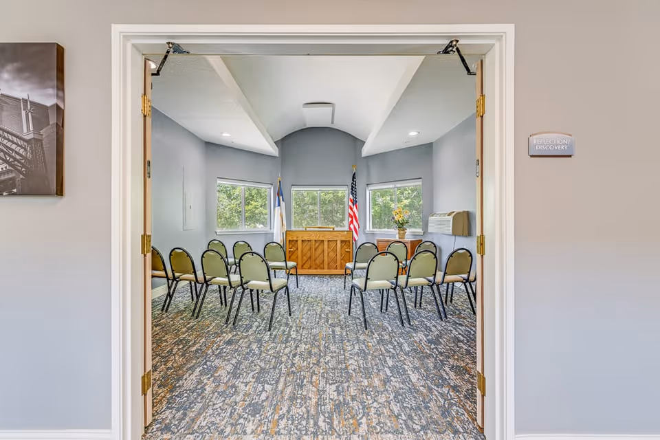 View through open double doors into a small meeting or chapel room with chairs arranged in rows facing a wooden piano and two flags near windows. The room has gray walls, a patterned carpet, and a sign on the wall outside the door that reads 'Reflection Discovery'.