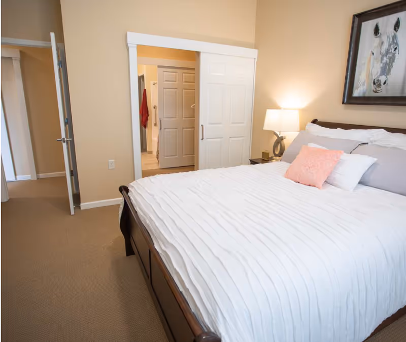 A bedroom with a large bed featuring white bedding and multiple pillows, including a pink decorative pillow. A bedside table with a lamp is next to the bed. The room has beige walls and carpeted floor. There is an open door leading to a hallway and a sliding door that opens to a bathroom with a visible red towel hanging.