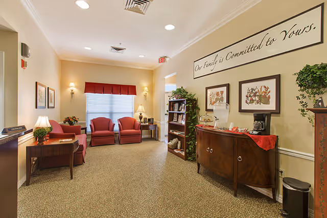 A cozy living room area in a senior living facility with beige walls and carpeted floor. The room features three red upholstered chairs arranged near a window with blinds and a red valance. There are two framed floral pictures on the wall, a wooden side table with a lamp and plant, a wooden cabinet with a water dispenser and coffee maker, and a bookshelf with various items. A sign above the cabinet reads 'Our Family is Committed to Yours.'