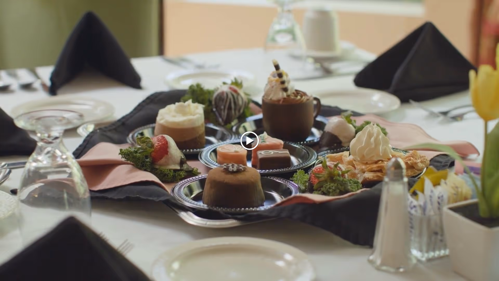 A close-up view of a dining table set with a variety of desserts on silver platters, including chocolate mousse, whipped cream-topped treats, strawberries, and other pastries. The table is covered with a white tablecloth and has black folded napkins, glassware, plates, and silverware arranged neatly. A yellow tulip in a small pot is visible on the right side of the table.