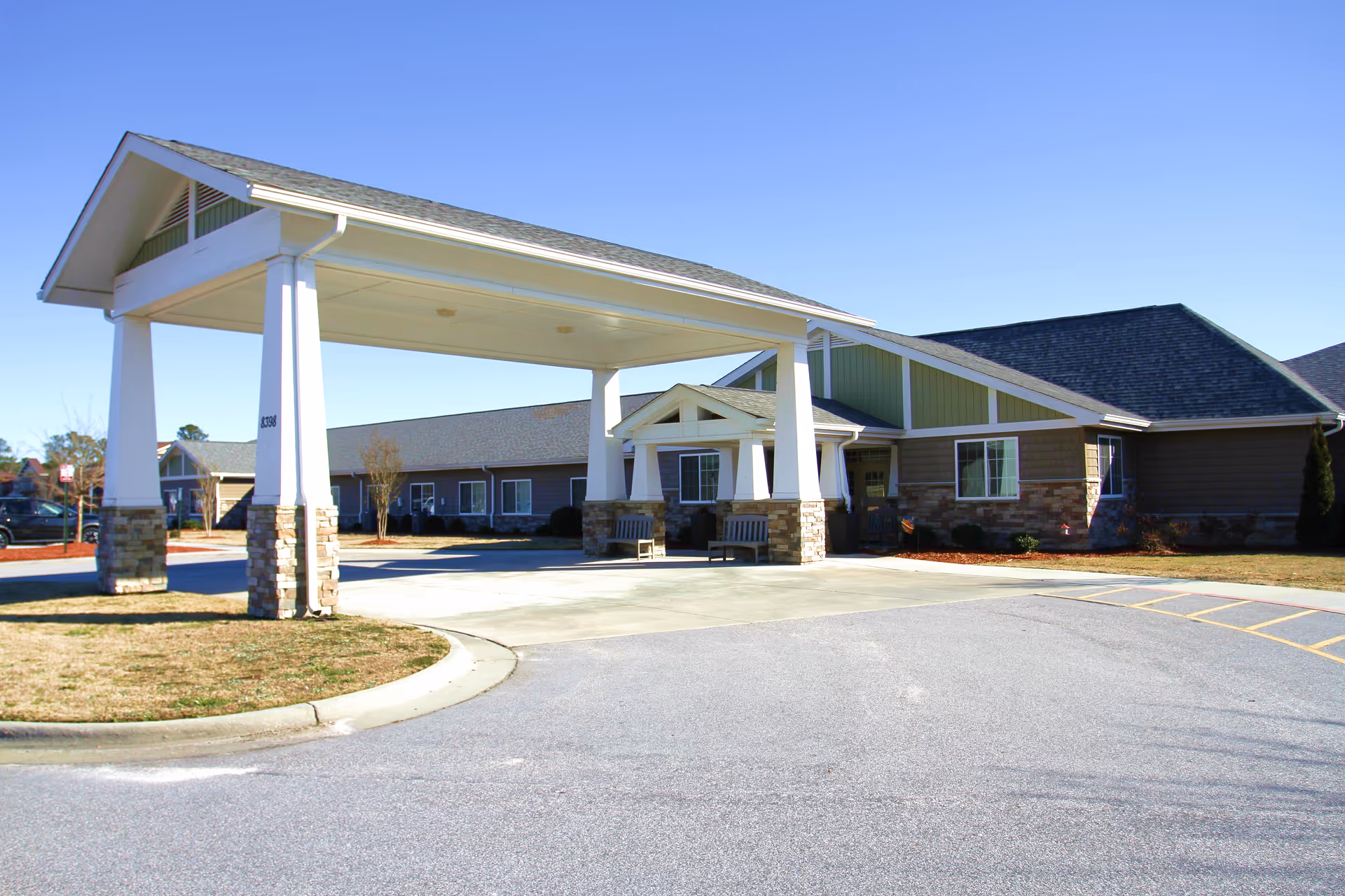 Entrance of a single-story senior living facility with a covered porte-cochere, benches, and a paved driveway.
