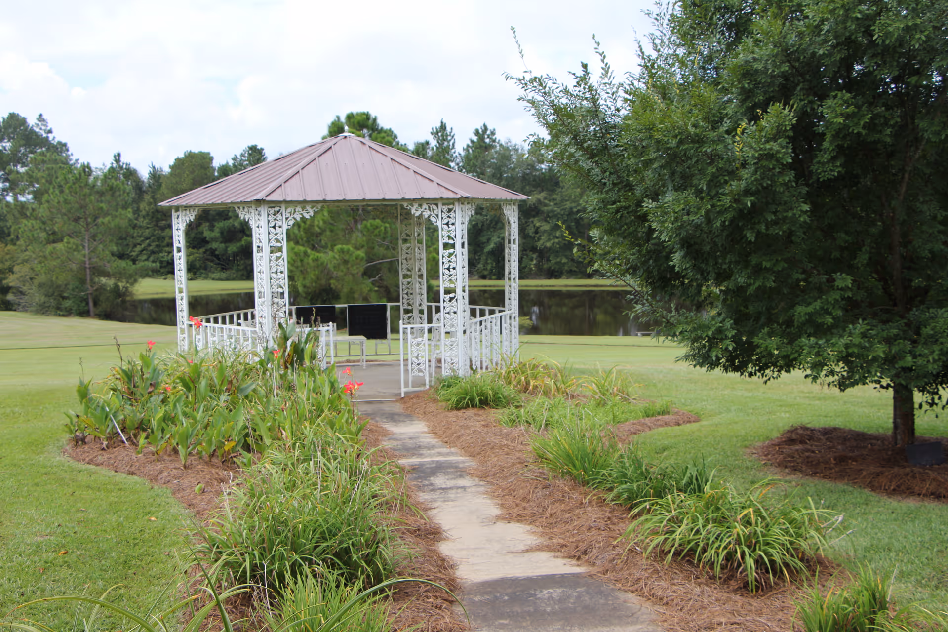 A white metal gazebo with a brown roof situated at the end of a paved pathway surrounded by green grass, plants, and trees, with a pond visible in the background under a partly cloudy sky.