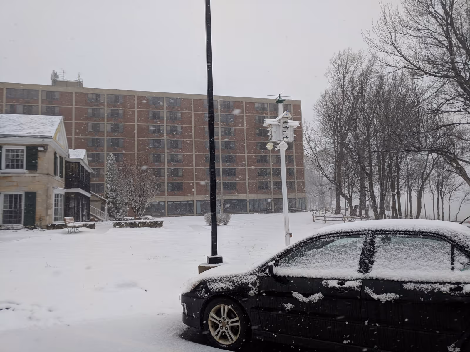Snow-covered outdoor scene at Brevillier Village showing a black car parked in the foreground with snow on its roof and windows. In the background, there is a multi-story brick building and a smaller house-like structure to the left. Bare trees and a birdhouse on a pole are also visible, with snow falling and covering the ground.