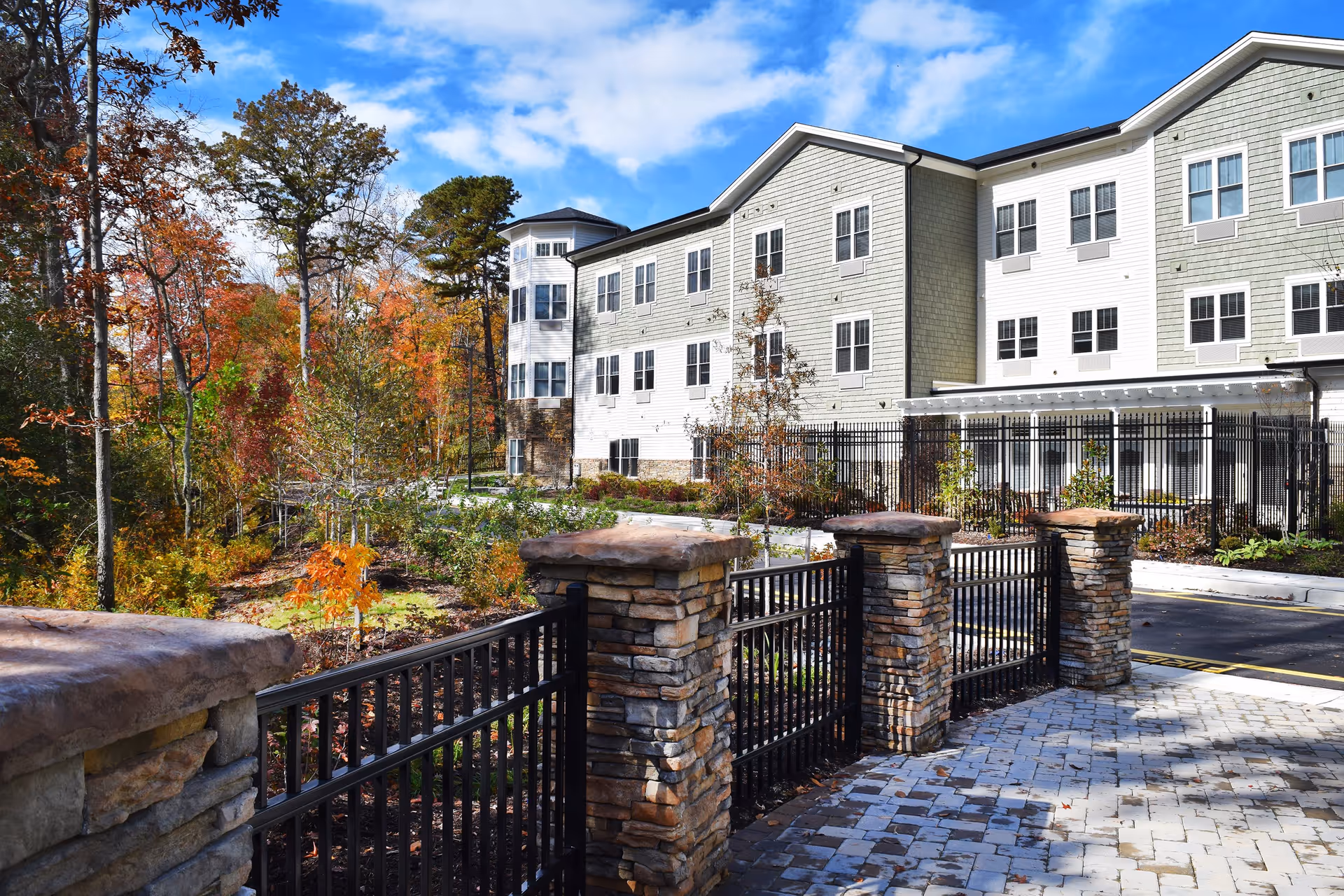 Exterior view of a multi-story senior living facility with light-colored siding and numerous windows, surrounded by autumn trees with colorful foliage. In the foreground, there is a stone and metal fence along a paved walkway under a partly cloudy blue sky.