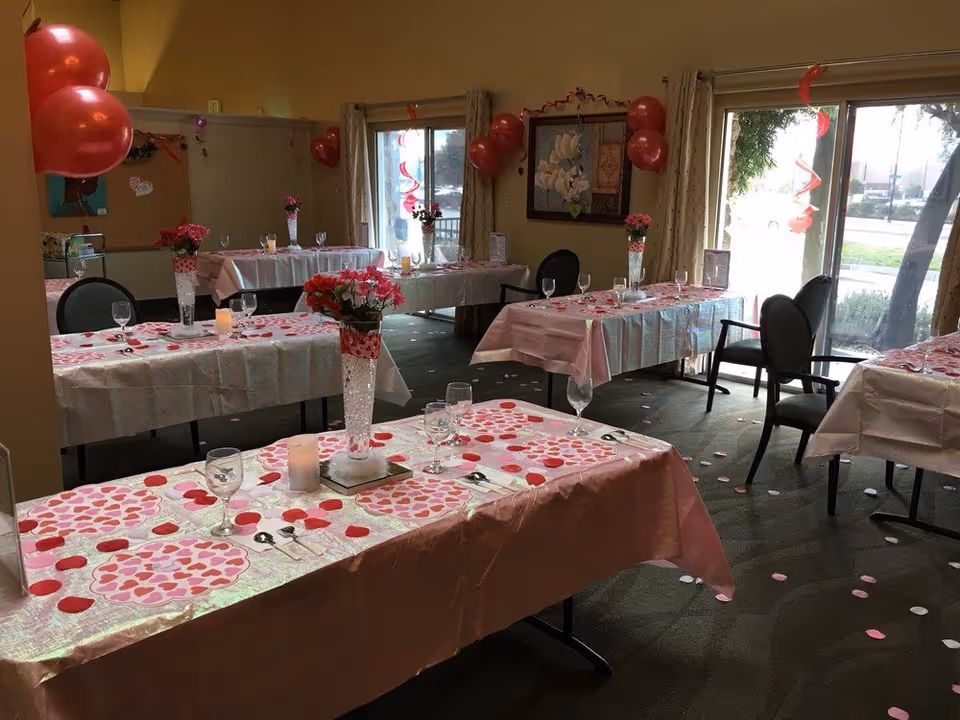 Decorated dining room with multiple tables covered in pink tablecloths, heart-shaped confetti, floral centerpieces and place settings.