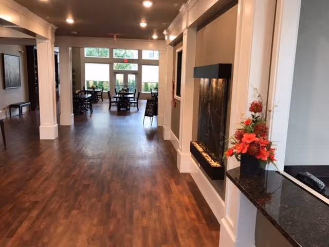 Interior view of a senior living facility hallway with wooden flooring, white pillars, a modern wall-mounted fireplace, a flower arrangement on a black countertop, and a dining area with tables and chairs near large windows at the far end.