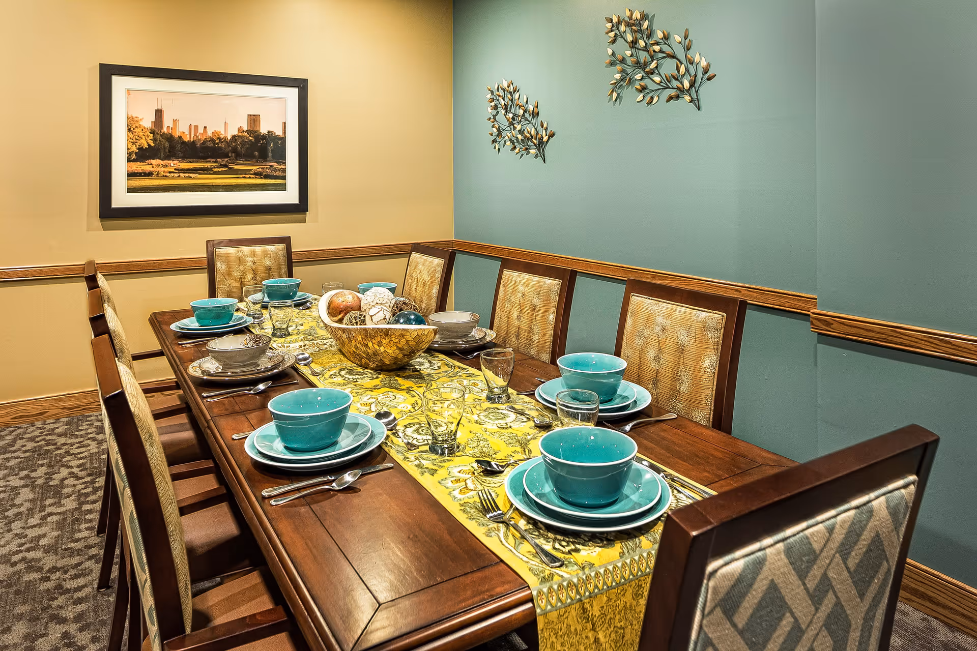 A dining room with a long wooden table set for eight people. The table has a yellow patterned runner and is set with turquoise bowls, plates, glasses, and silverware. The chairs have cushioned backs with a beige and brown pattern. The walls are painted in two tones: beige and teal, with decorative metal wall art and a framed picture of a cityscape on the beige wall.