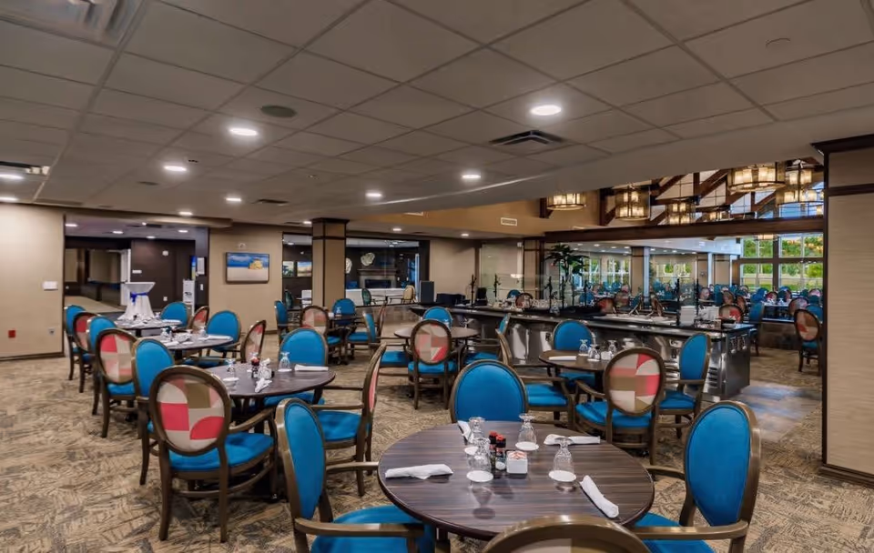 A spacious dining room in Seaside Springs Retirement Community with multiple round tables set with napkins, glasses, and condiments. The chairs have blue cushions and patterned backs. The room features a carpeted floor, a drop ceiling with recessed lighting, and large mirrors along one wall reflecting the seating area. There are windows providing natural light and decorative light fixtures hanging from the ceiling.