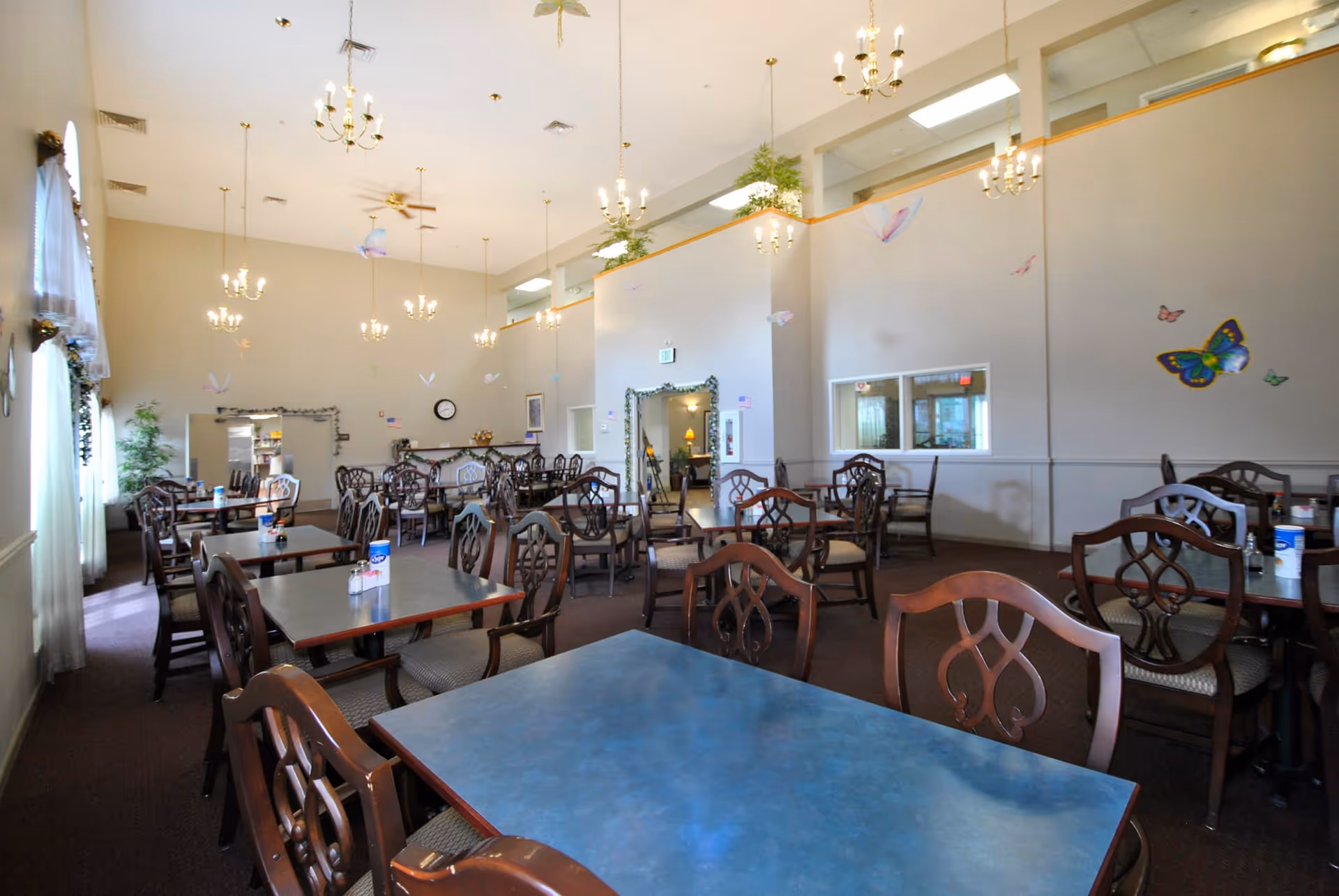 A spacious dining room with multiple dark wood tables and chairs arranged neatly. The room has high ceilings with several chandeliers hanging down, large windows with sheer curtains on the left side, and butterfly wall decorations on the right wall. There are plants and a clock on the far wall, and the room is well-lit with natural and artificial light.