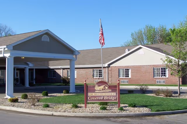 Exterior view of Covered Bridge Health Campus showing a single-story brick building with a covered entrance. An American flag is flying on a flagpole in front of the building, and there is a landscaped area with small bushes and rocks surrounding a sign that reads 'The Health Center at Covered Bridge.'