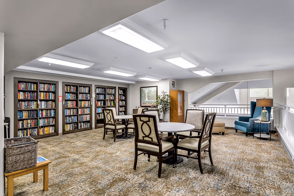 A bright and spacious library room with multiple bookshelves filled with books along the back wall. There are round tables with chairs arranged around them for reading or socializing. A cozy seating area with a blue armchair, ottoman, and side table with a lamp is visible near a window. The room has carpeted flooring and fluorescent ceiling lights.