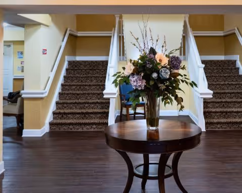 Interior view of a senior living facility showing a round wooden table with a large floral arrangement in a glass vase placed in the center. Behind the table, there are two carpeted staircases with white railings leading to an upper floor. The walls are painted in a warm beige color, and there is dark wood flooring.