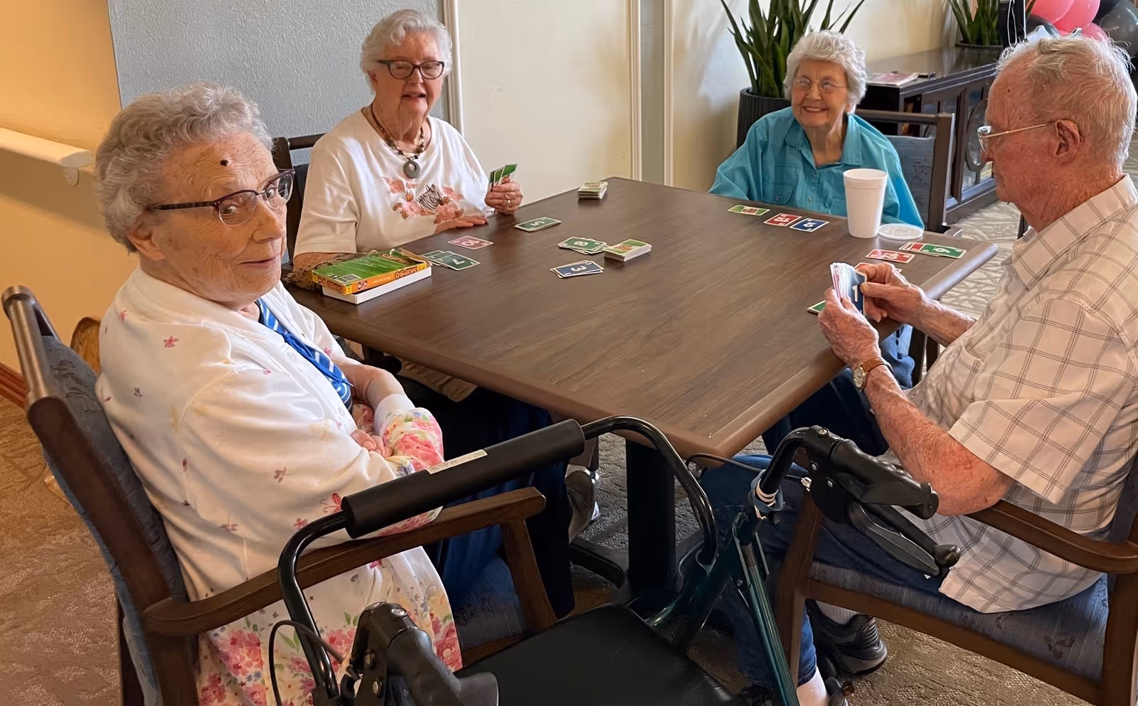Four elderly individuals sitting around a wooden table playing a card game in a well-lit room. Three women and one man are engaged in the game, with cards and a box of cards on the table. The man and one woman have walkers next to their chairs. There is a plant and a cabinet in the background.