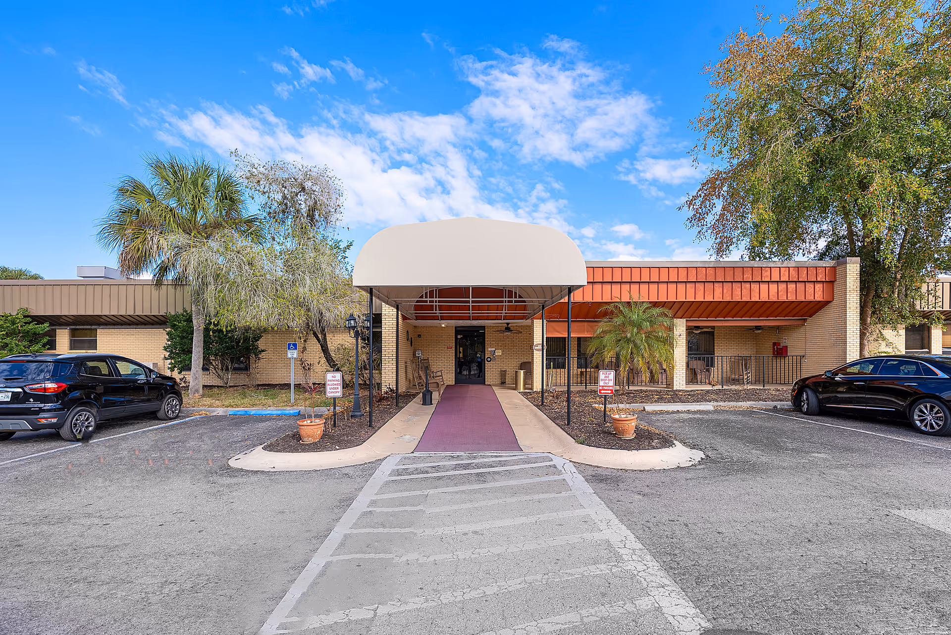 Front exterior view of North Port Rehabilitation and Nursing Center showing a single-story building with a covered entrance, a red carpet walkway, two potted plants, parked cars on either side, palm trees, and a partly cloudy blue sky.