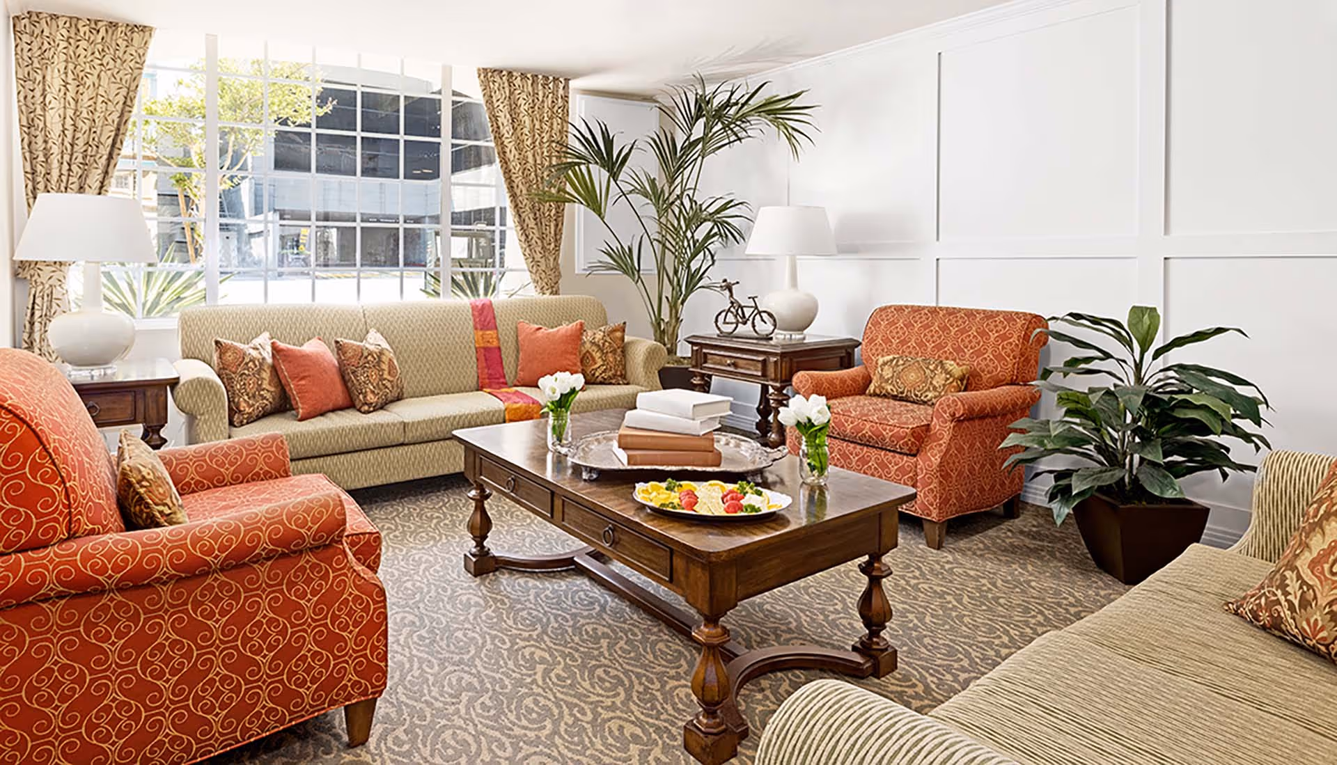 A bright and cozy living room in a senior living facility featuring two patterned red armchairs, two beige sofas with decorative pillows, a wooden coffee table with books and flower vases, large windows with patterned curtains, two table lamps, and green potted plants.