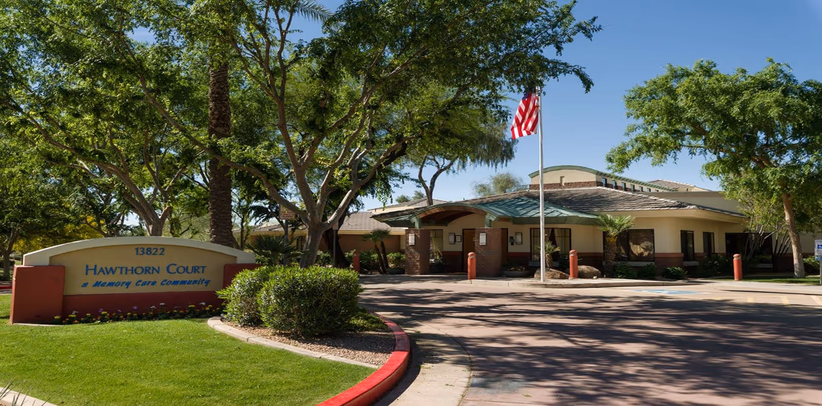 Exterior view of Hawthorn Court at Ahwatukee Memory Care facility with a sign displaying the name and address, surrounded by trees and greenery under a clear blue sky. An American flag is flying on a flagpole near the entrance.