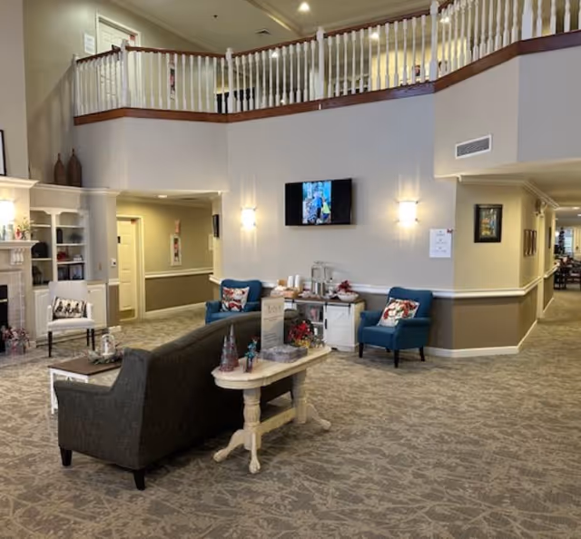 Spacious two-story senior living facility lobby with seating, a wall-mounted TV, decorative table, and balcony railing above.