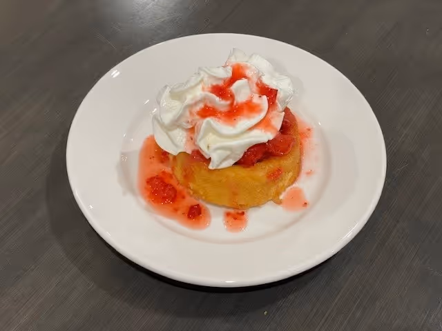 A small dessert cake topped with strawberry sauce and whipped cream on a white plate on a gray table.