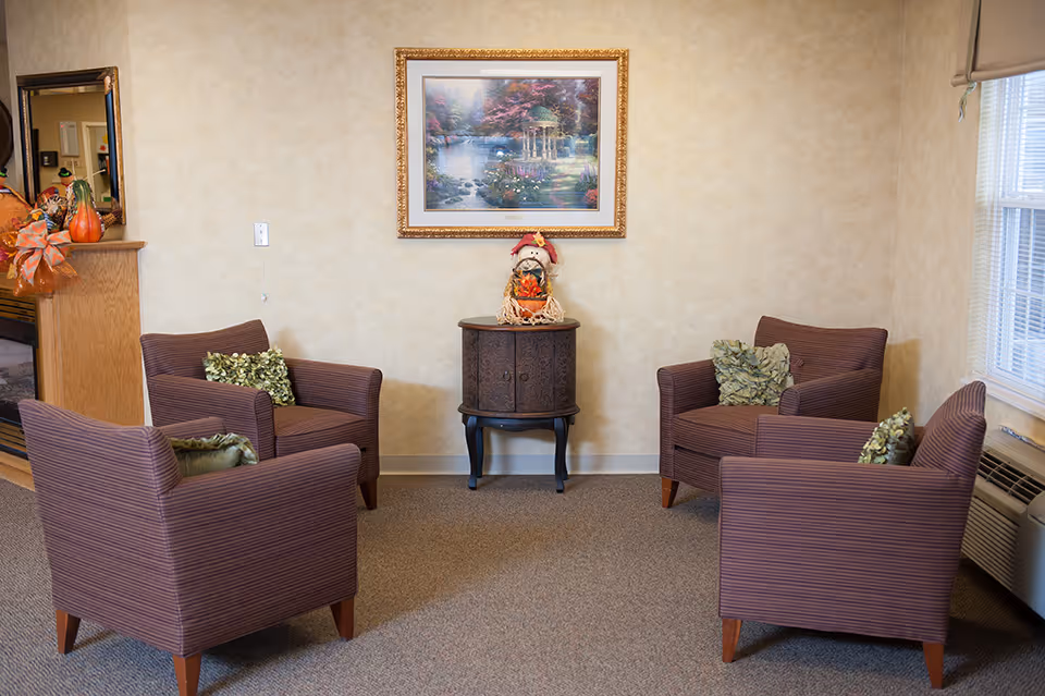 A cozy sitting area with four striped armchairs arranged in a circle around a small wooden cabinet. The cabinet has a decorative scarecrow figure on top. A framed painting of a garden scene hangs on the beige wall behind the cabinet. There is a window with blinds on the right side and a fireplace with autumn decorations on the left.
