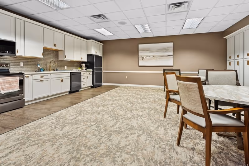 A spacious room featuring a modern kitchen area with white cabinets, a black refrigerator, stove, and dishwasher. The room also includes a dining area with wooden chairs and tables, beige walls, a large patterned carpet, and a framed abstract painting on the wall.