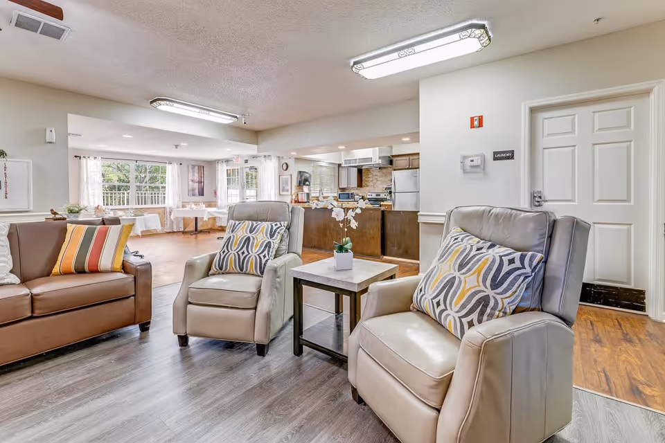 A bright and spacious senior living common area featuring two beige leather armchairs with patterned cushions, a brown leather sofa with striped cushions, a small table with a white orchid plant, and a kitchen area in the background. Large windows with sheer curtains allow natural light to fill the room.