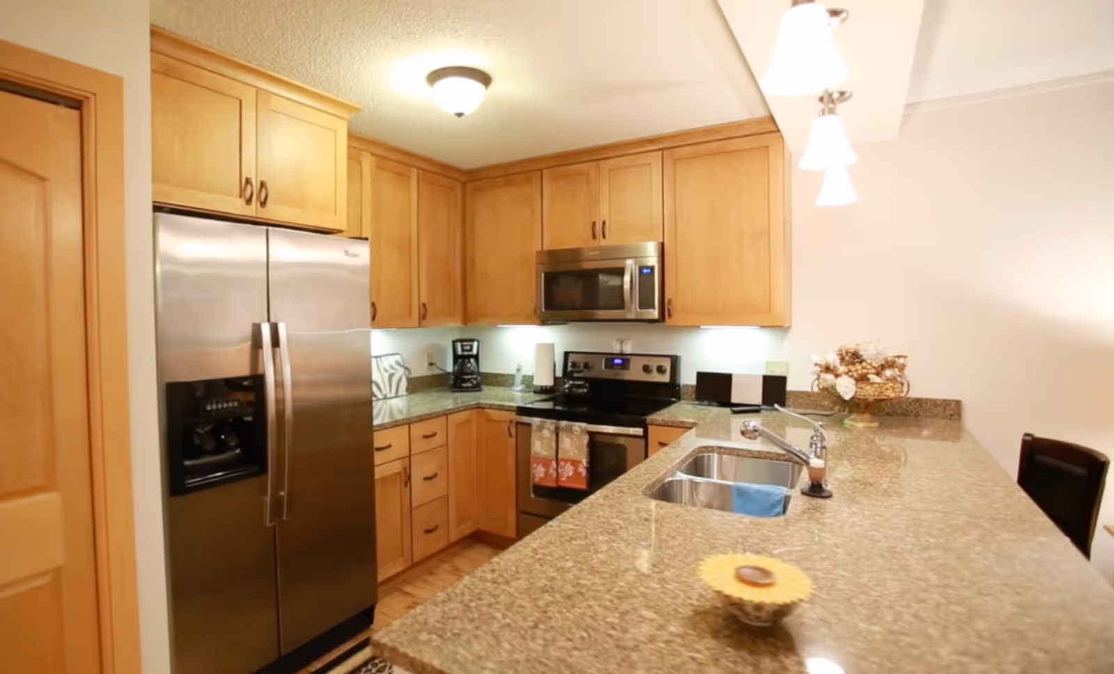 A modern kitchen with wooden cabinets, a stainless steel refrigerator, microwave, and stove. The kitchen has granite countertops with a sink and faucet, a coffee maker, and decorative items on the counter.