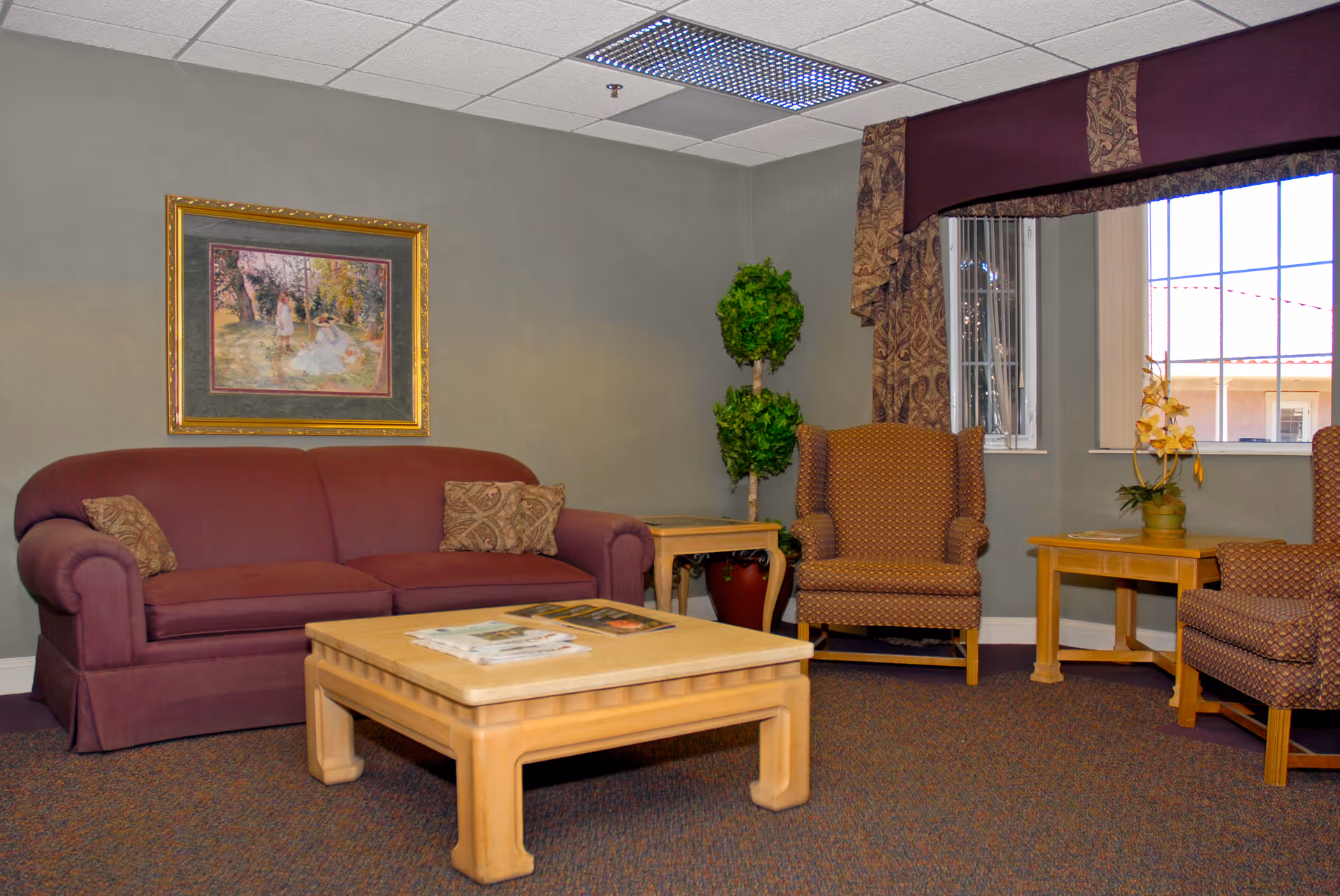 A cozy living room area with a maroon sofa, two patterned armchairs, a light wood coffee table with magazines on it, a side table with a potted plant, and a framed painting on the wall. There is a window with decorative curtains letting in natural light.