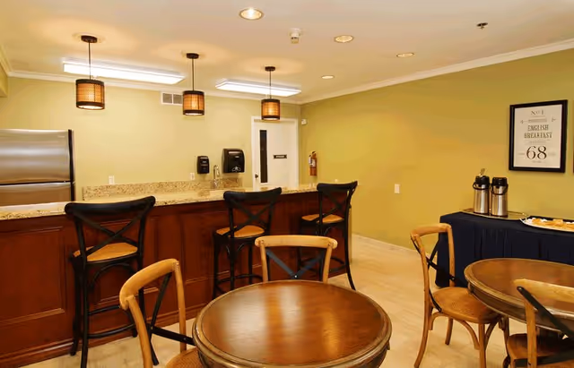 Interior view of a dining area with round wooden tables and chairs. There is a counter with three high chairs, pendant lights hanging above, and a stainless steel refrigerator. On the right side, a table with coffee dispensers and plates is visible against a light green wall with a framed print.