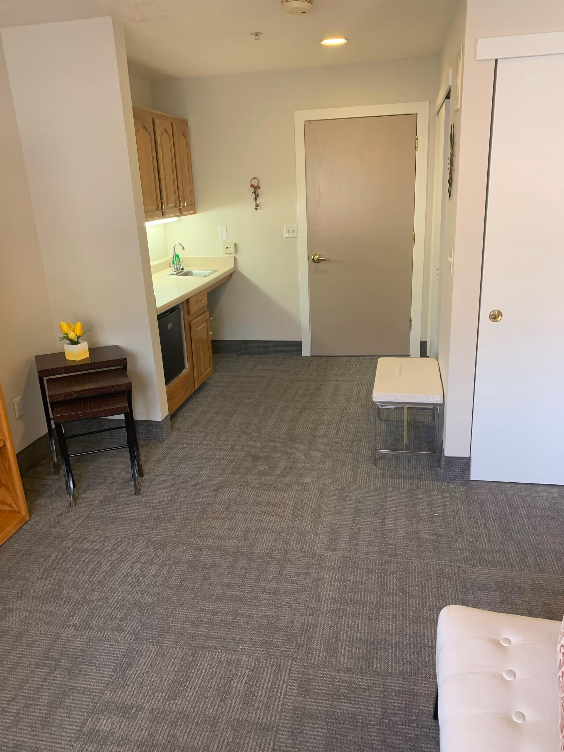 Interior view of a room in Ridgeview Gardens Assisted Living featuring a small kitchenette with wooden cabinets, a sink, and a mini fridge. There is a closed door in the background, a small cushioned bench on the right, and a set of nested tables with a small vase of yellow flowers on the left. The floor is carpeted with a gray patterned carpet.