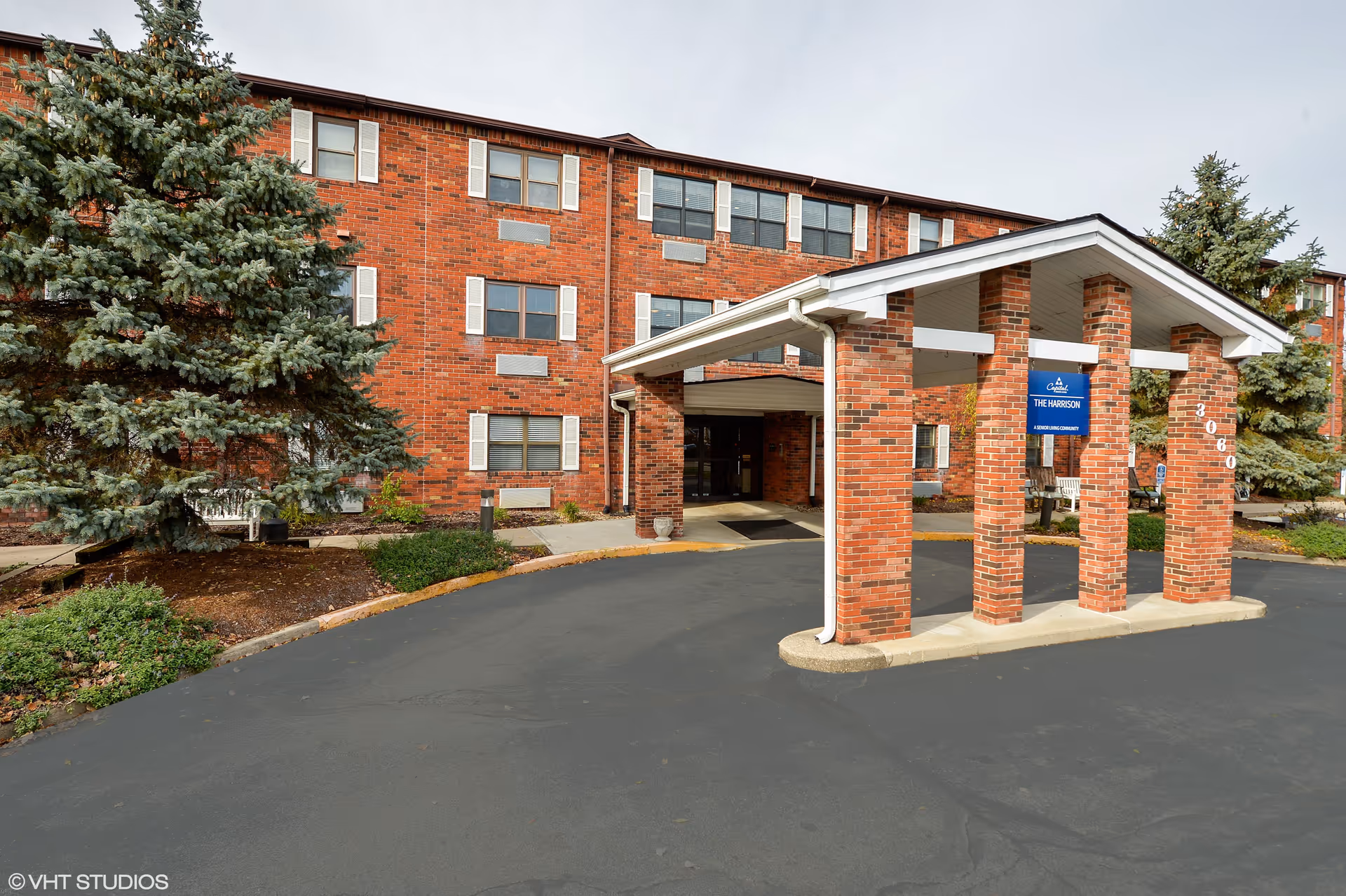 Exterior view of The Harrison senior living facility showing a red brick building with multiple windows and white shutters. There is a covered entrance supported by brick columns with a blue sign that reads 'The Harrison'. The area is surrounded by greenery including large evergreen trees and shrubs.
