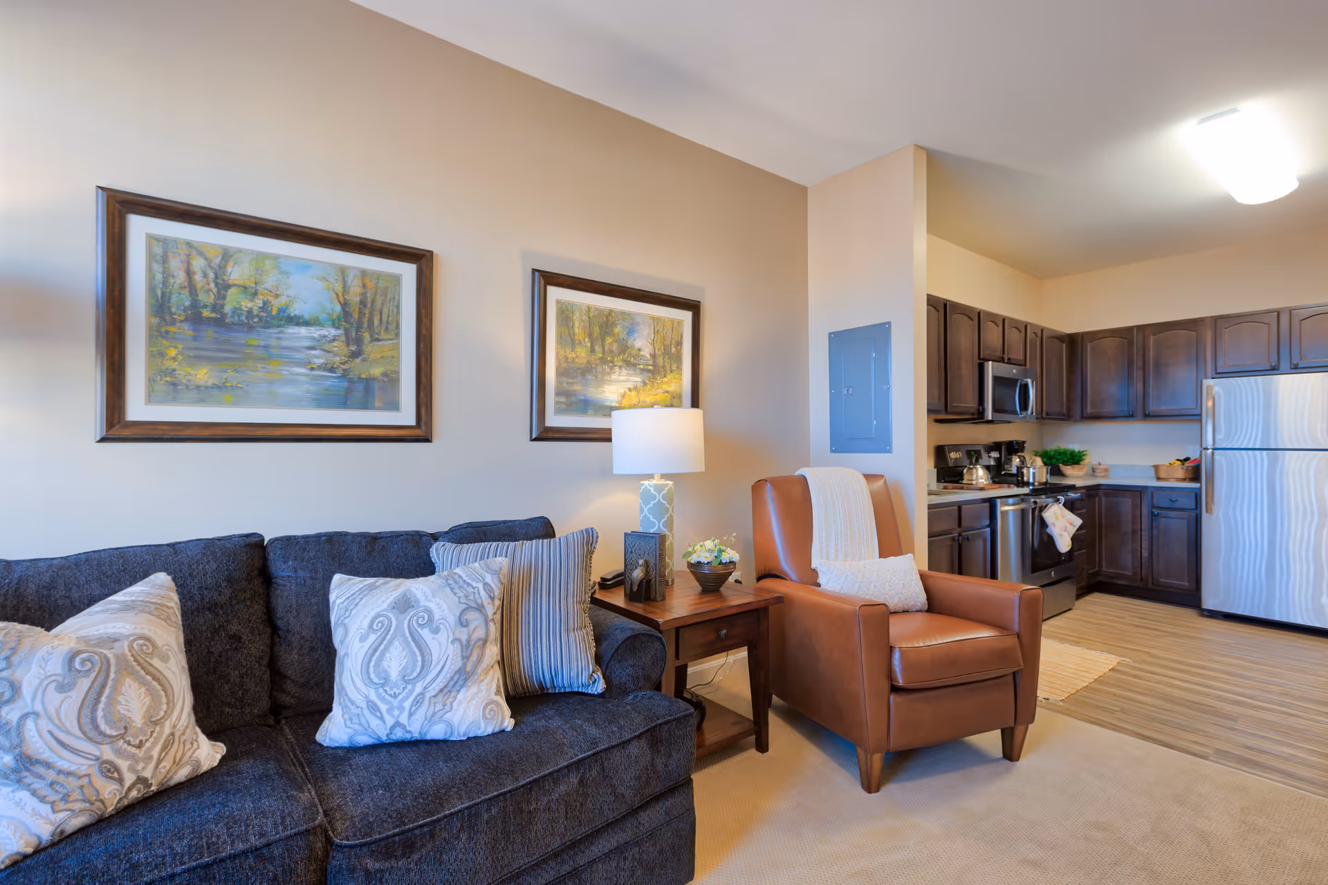 A cozy living area with a dark gray sofa adorned with patterned pillows, a brown leather armchair with a white throw blanket, and a wooden side table with a lamp and decorative items. The background shows a kitchen with dark wood cabinets, stainless steel appliances including a refrigerator, microwave, and stove, and a light wood floor. Two framed landscape paintings hang on the beige wall above the sofa.