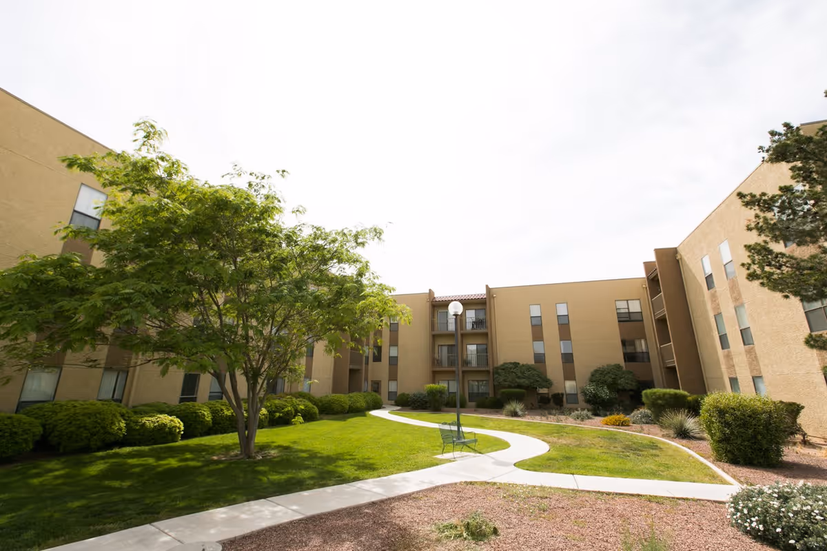 Outdoor courtyard area of a senior living facility with a winding concrete pathway, green grass, bushes, trees, and a bench under a lamp post. The courtyard is surrounded by a three-story beige building with multiple windows and balconies.