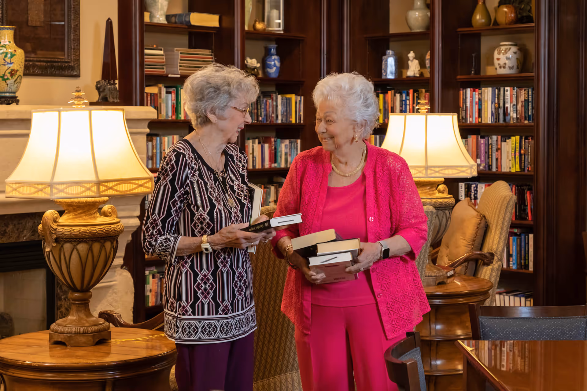 Two elderly women standing and holding books in a cozy library room with wooden bookshelves filled with books and decorative items. The room is warmly lit by two table lamps on wooden side tables, and there are comfortable chairs nearby.