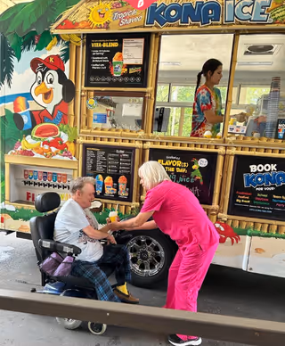 An elderly man in a wheelchair is being assisted by a woman in bright pink scrubs in front of a colorful Kona Ice shaved ice truck. The truck features a tropical theme with a cartoon penguin mascot and a menu of shaved ice flavors. A person inside the truck is preparing orders.